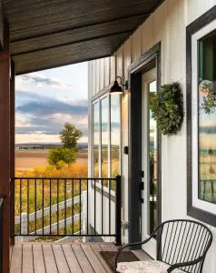 Porch entrance to a granny flat. Covered wooden porch, black railing, looking down the side of the house and out into a golden field with a Colorado sunset in the sky. The house has white siding, with dark grey framing around a white glass door and windows.