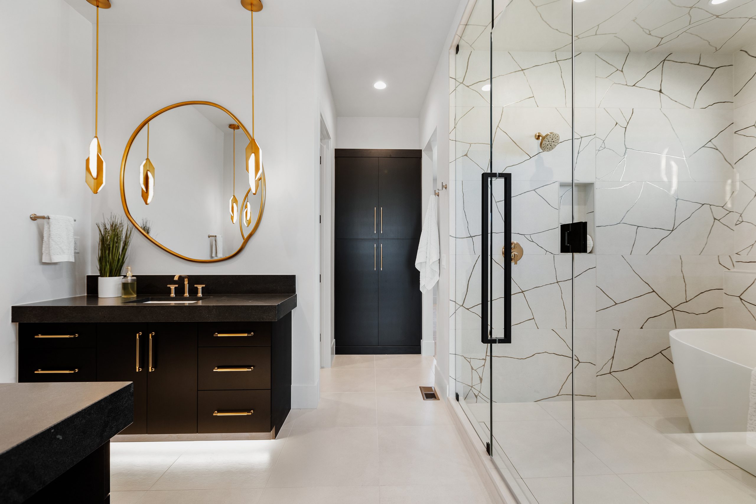 Marble walled shower, deep soaker tub, dark dual fantasies, with gold handles. There are gold pendants hanging down, with a mirror that this round and golden framed. The shower and tub are large and encased with glass, with long black handles. Down a hallway there is large dark cabinet with long gold handles. A luxury bathroom.