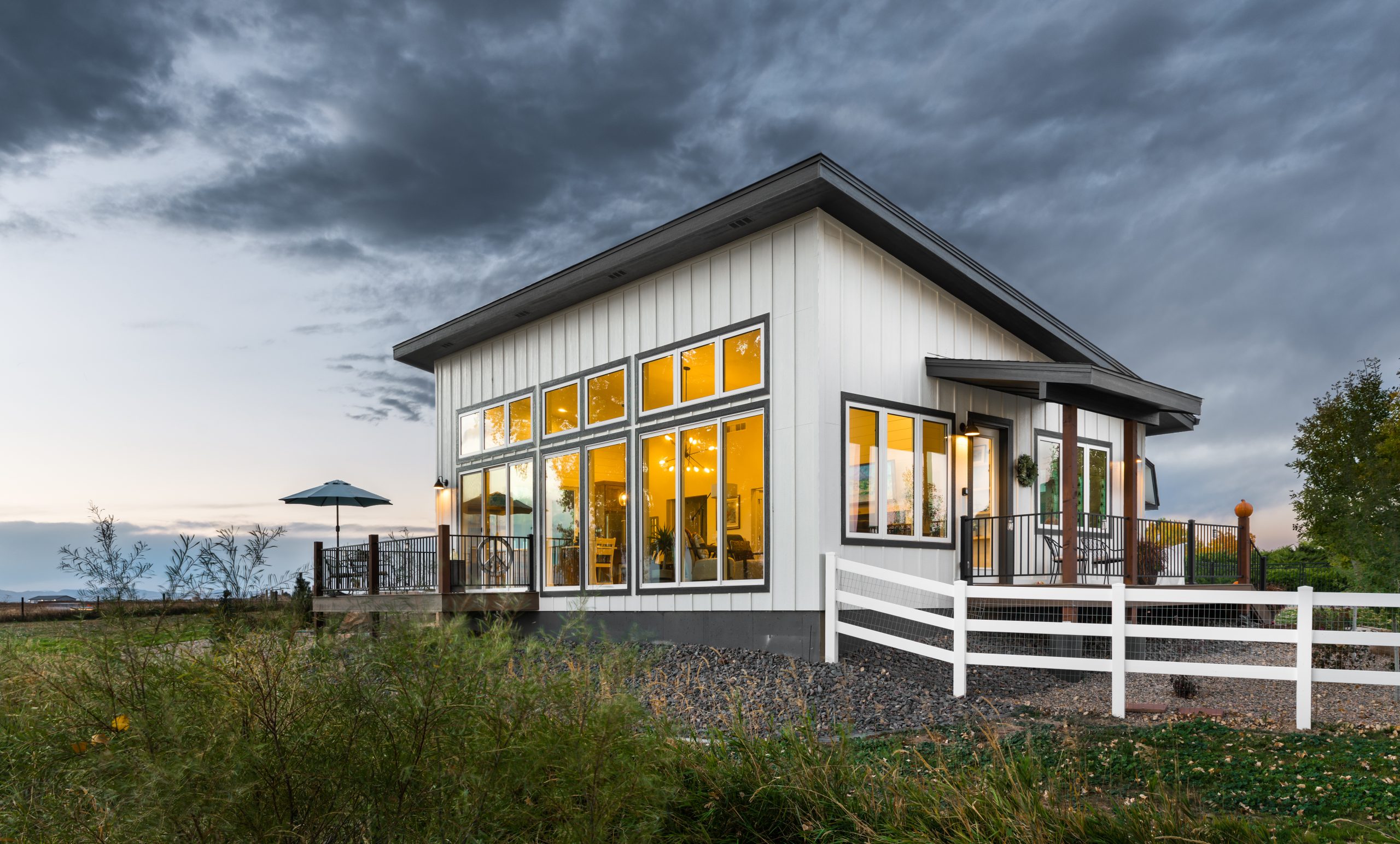 A granny flat, built in Colorado. The sky is dark and there are some storm clouds during the sunset. There is white fencing off of one side of the house, white in the front side shown is floor to ceiling windows. The windows glow with warm light inviting you in.