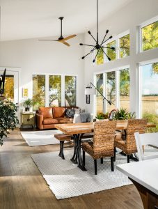 Open granny flat floor plan, with dinning space, and in the back couch seating. There is a fan on the ceiling, and a modern black chandelier for lighting. On one wall there is a black modern fireplace, and on all other surrounding walls are floor to ceiling windows. 