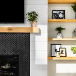 A herringbone black tiled fireplace, with a light wooden beam as a mantle. There is white shiplap on the wall, and off to the right is a build in book case. The shelves are all light wood matching the mantle.