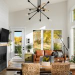 Full view of the living room, with a glass door on the left, and 3 large window panels on the right. A black modern chandelier drops down from the ceiling, and a tan couch fits perfectly under the windows. Off to the lefts is a fire place and tv that hangs from the wall.