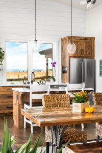 Wooden cabinetry on the back wall, framing a large window over looking the mountains and a field, placed right above the kitchen sink. There is a wooden island with white counter tops all through out the kitchen. Above the kitchen island are two drop down glass lights. Closer is a wooden table, with wicker chairs.