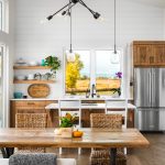 White shiplap walls, brown cabinetry sit above the fridge, and against the far back walls. There is white counter tops lining the back wall with the kitchen sink, and that same countertop can be seen on a kitchen island in front of the sink. There is a large kitchen window above the sink, that opens up the space and lights the area. Off to the left is a set of 2 floating shelves in a wood color that matched the cabinetry.