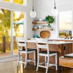 Full of light and color, there are floor to ceiling windows on the left side of the photo, where you see trees and railing from a porch. On the right are two white bar top chairs lining a wooden kitchen island with white counter tops. In the center of the photo you see two wooden floating shelves, and above that are two drop down glass lights above the kitchen island.