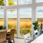 This is a photo of the floor to ceiling windows, overlooking golden fields in Colorado. in the bottom right corner there is a set of white chair tops lined by the white counter top. On the bottom left corner there are a set of wicker chairs tucked under a wooden kitchen table.